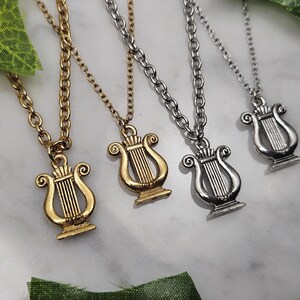 Puede incluir: Cuatro collares con colgantes de lira. Dos son dorados y dos plateados. Los colgantes presentan líneas verticales y adornos decorativos. Las cadenas están enlazadas y descansan sobre una superficie de mármol blanco.