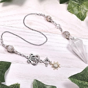 May include: A white crystal pendulum with a silver chain and decorative charms, including a lyre, arrow, and sun. The pendulum is hanging against a white wooden background with green leaves.