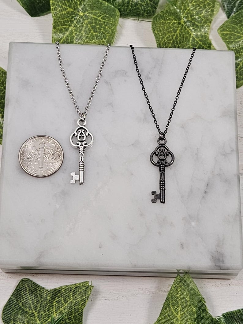 Puede incluir: Dos collares con llaves de plata, uno con una cadena negra y otro con una cadena plateada. Las llaves son ornamentadas y tienen un aspecto vintage.