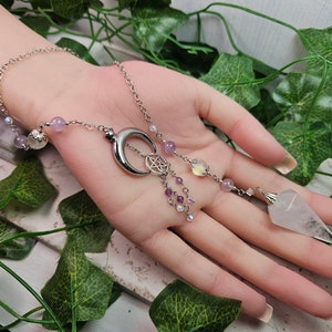 May include: A silver chain necklace with a crescent moon pendant and a pentagram charm. The necklace is adorned with purple and clear beads and a clear crystal pendulum.