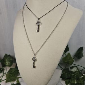 Puede incluir: Dos collares de plata con forma de llave superpuestos en un maniqu&iacute; blanco. El collar superior tiene una cadena negra y el collar inferior tiene una cadena plateada. Ambos collares presentan un colgante de llave de plata.