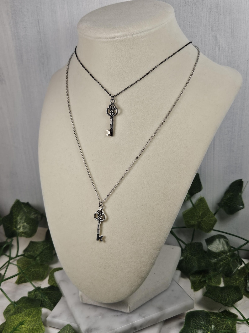 Puede incluir: Dos collares de plata con forma de llave superpuestos en un maniqu&iacute; blanco. El collar superior tiene una cadena negra y el collar inferior tiene una cadena plateada. Ambos collares presentan un colgante de llave de plata.