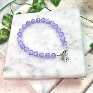 Puede incluir: Un brazalete de cuentas de color lavanda con un dije de mariposa plateado. El brazalete está sobre una superficie de mármol blanco.