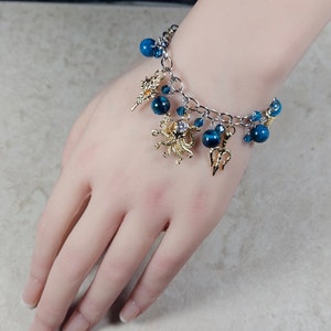 May include: A silver chain bracelet with blue beads and gold charms. The charms include a seashell, an octopus, and a trident. The bracelet is adorned with small blue crystals and is displayed on a person's hand.