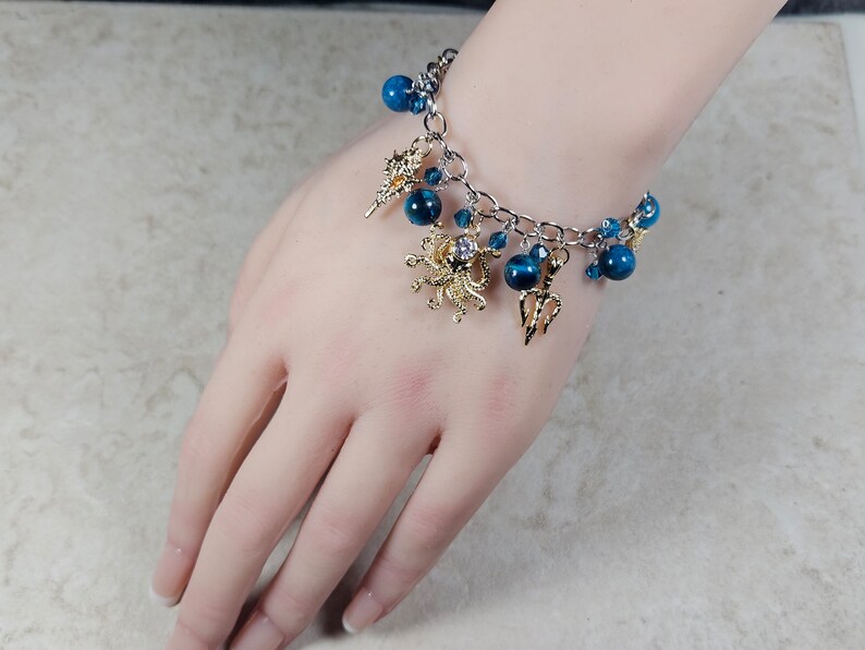 May include: A silver chain bracelet with blue beads and gold charms. The charms include a seashell, an octopus, and a trident. The bracelet is adorned with small blue crystals and is displayed on a person's hand.