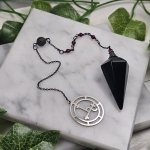 May include: Black obsidian pendulum with a silver chain and a silver charm featuring a symbol. The pendulum is hanging from a white surface.