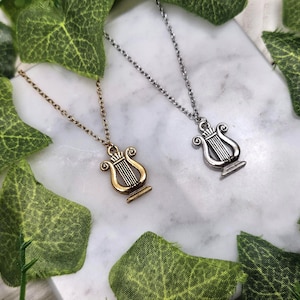 Op de afbeelding: Twee zilveren kettingen met een lier hanger. De lier is een muziekinstrument met snaren. De kettingen liggen op een witte achtergrond met groene bladeren.