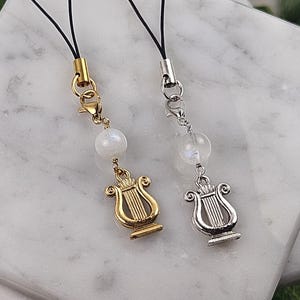 May include: Two decorative charms with a musical theme. One charm is gold-colored, the other is silver-colored. Each charm features a lyre pendant, a white bead, and a black cord with a metal clasp. The charms are designed to be attached to a phone or other device.