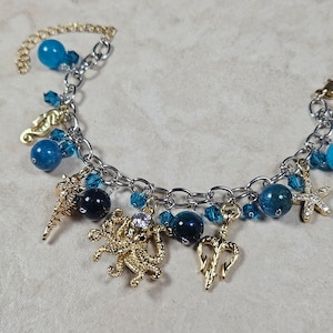 Puede incluir: Una pulsera de dijes plateada con dijes temáticos del océano. La pulsera incluye dijes dorados de caballito de mar, estrella de mar y tridente, junto con cuentas azules y azul oscuro. Una cadena dorada se extiende desde la pulsera.