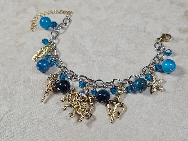 May include: A silver-toned charm bracelet featuring ocean-themed charms. The bracelet includes gold-toned seahorse, starfish, and trident charms, along with blue and dark blue beads. A gold-toned chain extends from the bracelet.
