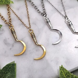 May include: Four necklaces with crescent moon pendants. Two are gold-toned, and two are silver-toned. Each pendant has a detailed handle design and hangs from a matching chain. The necklaces are displayed on a white marble surface.