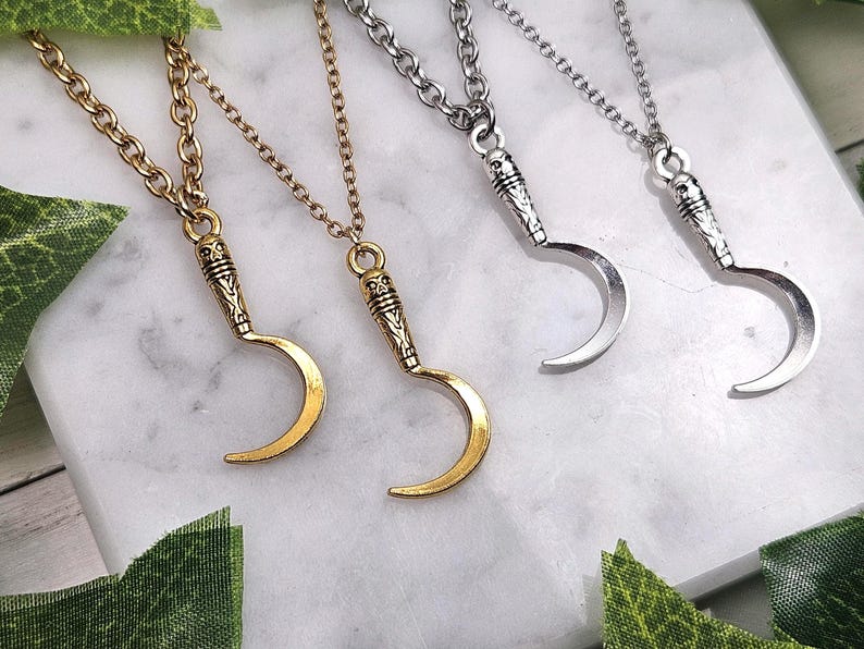 May include: Four necklaces with crescent moon pendants. Two are gold-toned, and two are silver-toned. Each pendant has a detailed handle design and hangs from a matching chain. The necklaces are displayed on a white marble surface.