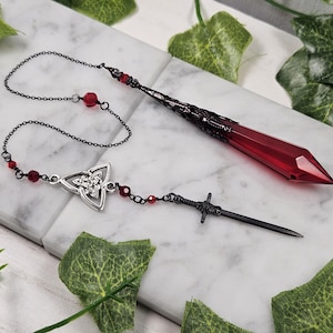 May include: A black chain necklace with a red crystal pendant and a silver Celtic knot charm. The necklace also features a black metal sword charm.