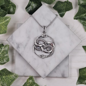 May include: A silver necklace with a pendant featuring two intertwined snakes forming an infinity symbol. The necklace is on a white marble surface.