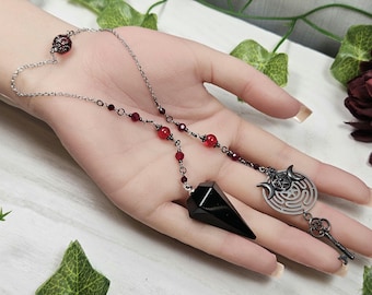 May include: A black crystal pendulum with a silver chain and red beads. The pendulum is attached to a silver charm with a pentagram and crescent moons.