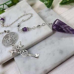 Può includere: Pendolo di ametista con una catena e ciondoli d'argento, tra cui una chiave, una luna crescente e un pentagramma. I ciondoli sono attaccati a un cerchio d'argento con un design a labirinto.