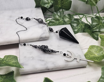 Puede incluir: Un péndulo con una punta de piedra negra pulida unida a una cadena de metal oscuro. La cadena está adornada con cuentas texturizadas negras y plateadas y un dije de metal plateado con forma de media luna combinada con un símbolo de cruz.