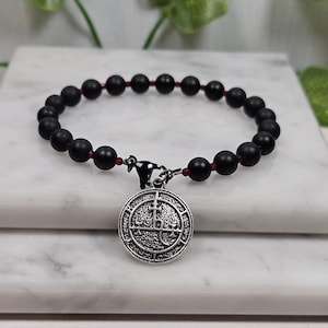 Puede incluir: Pulsera de cuentas negras con un dije de plata con un símbolo. La pulsera tiene detalles de cuerda roja.