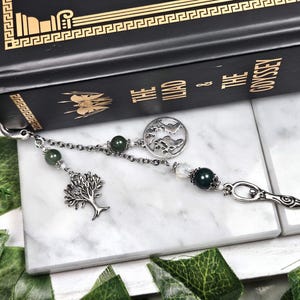 May include: A silver bookmark with a tree charm, green beads, and a circular charm with a mountain design. The bookmark is next to a black book titled "The Iliad & The Odyssey".