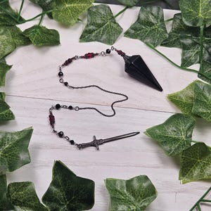 May include: A black obsidian pendulum with a silver chain and a silver sword charm. The pendulum is surrounded by green leaves.
