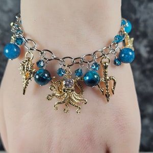 May include: A silver-toned charm bracelet featuring blue beads and various gold-colored charms. Charms include a seashell, octopus, and trident. Additional blue crystal accents add sparkle to the bracelet.