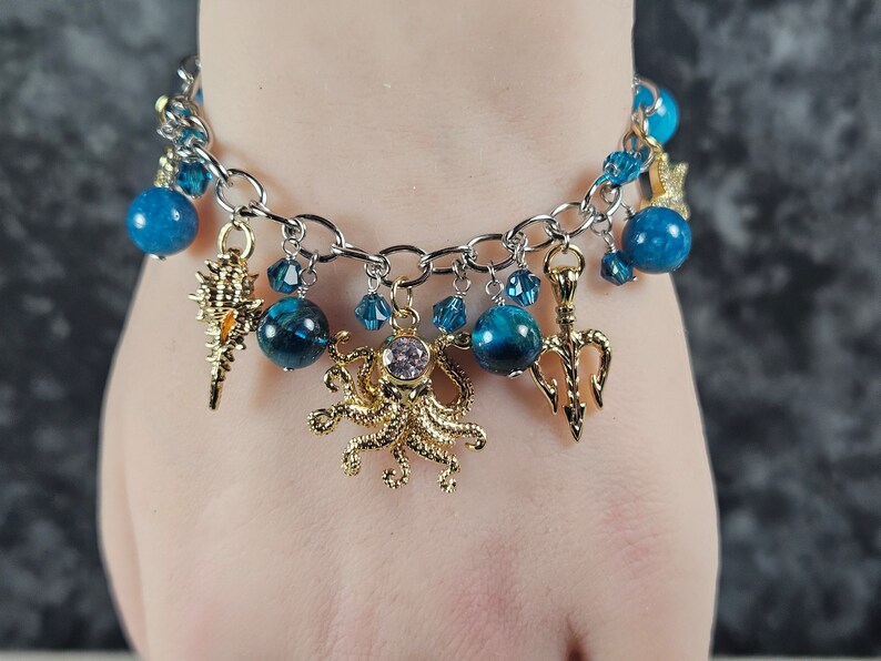 May include: A silver-toned charm bracelet featuring blue beads and various gold-colored charms. Charms include a seashell, octopus, and trident. Additional blue crystal accents add sparkle to the bracelet.