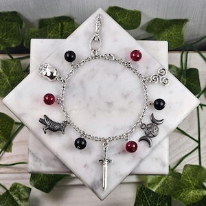 Peut inclure: Bracelet à breloques argenté avec des breloques en forme d'épée, de corbeau, de crâne et de croissant de lune avec un pentagramme, ainsi que des perles noires et rouges. Le bracelet est présenté sur une surface en marbre blanc.