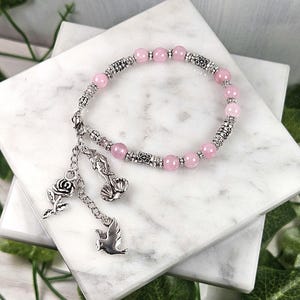 Puede incluir: Una pulsera de cuentas rosas con detalles y abalorios plateados. La pulsera presenta cuentas rosas redondas y cuentas plateadas decorativas. Los abalorios incluyen una rosa, una concha y un pájaro. La pulsera se exhibe sobre una superficie de mármol blanco.