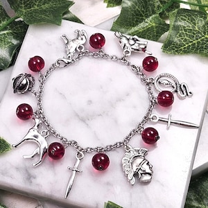 Puede incluir: Un brazalete de plata con cuentas de vidrio rojas y varios dijes de plata, incluyendo una serpiente, una espada, una corona, un lobo y un casco de guerrero.