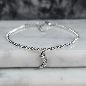 Peut inclure: Un bracelet en argent avec un petit charme en forme de croissant de lune. Le charme est serti de petites pierres claires.