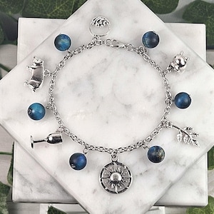 May include: A silver charm bracelet featuring a variety of charms, including a bear, a wine glass, a sunburst, and a leaf. The bracelet also has blue beaded accents. The bracelet is displayed on a white marble surface.
