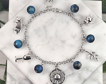 May include: A silver charm bracelet featuring a variety of charms, including a bear, a wine glass, a sunburst, and a leaf. The bracelet also has blue beaded accents. The bracelet is displayed on a white marble surface.