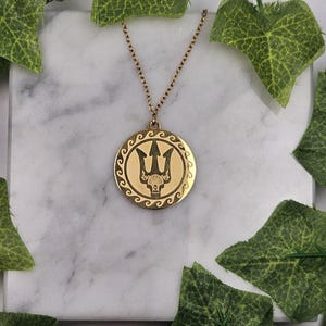 May include: A gold necklace with a round pendant featuring a trident design. The pendant has a wave pattern around the edge.