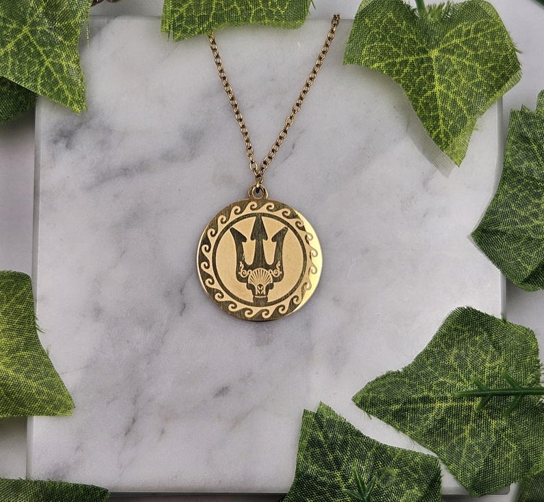 May include: A gold necklace with a round pendant featuring a trident design. The pendant has a wave pattern around the edge.