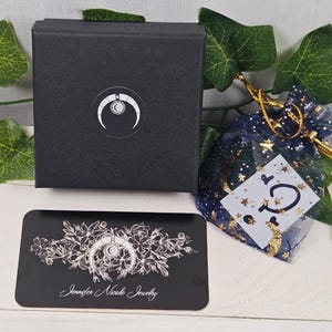 May include: A black box with a white crescent moon logo and the text "Jennifer Nicole Jewelry" on a black card. The box is sitting on a white surface with a blue and gold star-shaped pouch.