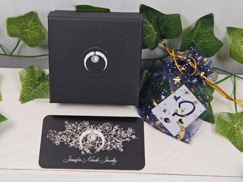 May include: A black box with a white crescent moon logo and the text "Jennifer Nicole Jewelry" on a black card. The box is sitting on a white surface with a blue and gold star-shaped pouch.
