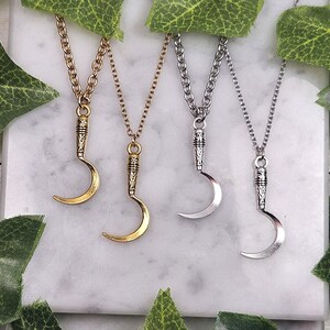 May include: Four necklaces with crescent moon pendants. Two are gold-toned, and two are silver-toned. Each pendant is suspended from a chain. The necklaces are displayed on a white marble surface, with green leaves in the background.