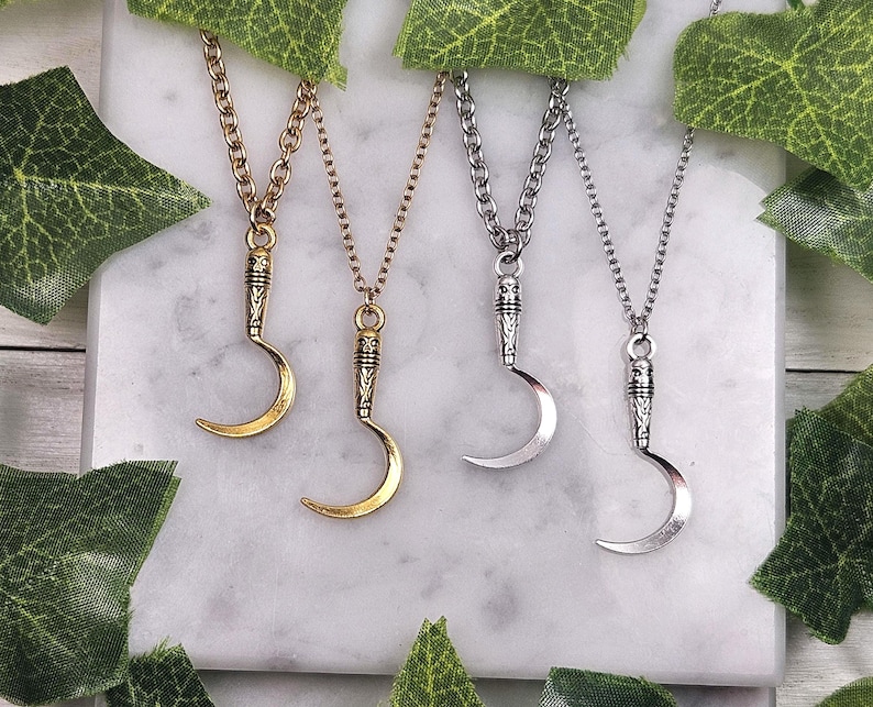May include: Four necklaces with crescent moon pendants. Two are gold-toned, and two are silver-toned. Each pendant is suspended from a chain. The necklaces are displayed on a white marble surface, with green leaves in the background.