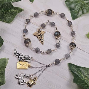 Könnte beinhalten: Eine graue und goldene Perlenkette mit verschiedenen Anhängern, darunter ein medizinisches Symbol, ein geflügelter Schuh, eine Schildkröte und ein Hahn. Die Kette ist aus Labradorit-Steinen gefertigt.