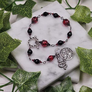 Pode incluir: Uma pulseira de contas pretas e vermelhas, conectadas por uma corrente prateada. Possui um fecho de alternância e um pingente com uma cobra e um emblema circular. Folhas verdes circundam a pulseira.