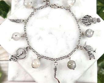 May include: A silver charm bracelet with multiple charms, including a crescent moon, a snake, a bird, and wings. The bracelet also features several round, iridescent stones.