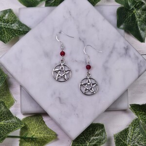 May include: A pair of silver earrings with a pentagram charm and a red crystal bead on each earring.