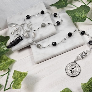 May include: A necklace featuring black and grey beads, clear crystal beads, and a silver pendant. The pendant has a circular design with a stylized symbol. A black crystal and silver charms hang from the necklace.