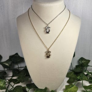 Op de afbeelding: Twee gouden kettingen met een lier hanger. De bovenste ketting is zilver en de onderste ketting is goud.