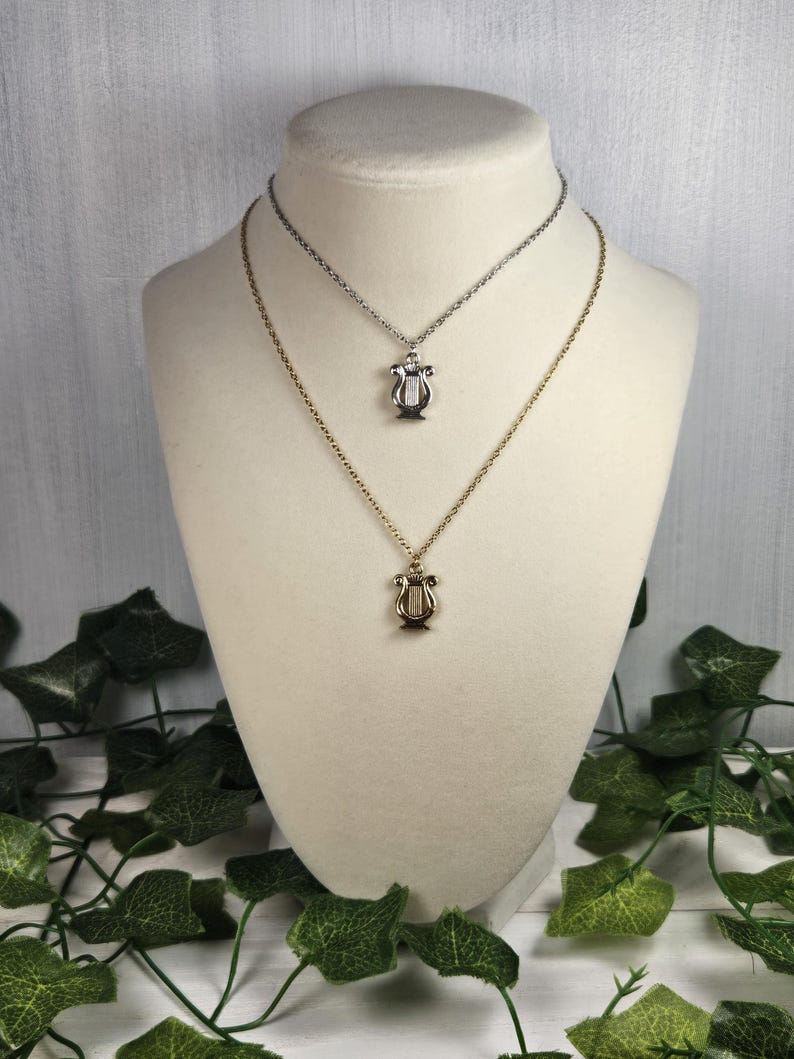 Op de afbeelding: Twee gouden kettingen met een lier hanger. De bovenste ketting is zilver en de onderste ketting is goud.