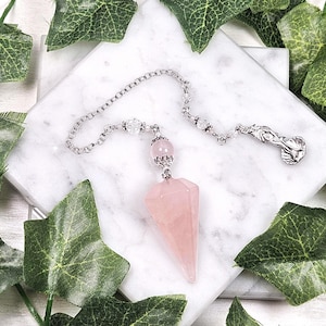 Pode incluir: Um pêndulo de quartzo rosa rosa com uma corrente de prata e um amuleto de prata. O pêndulo está sobre uma superfície branca com folhas verdes.