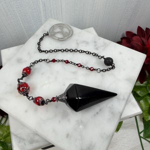 Könnte beinhalten: Ein schwarzes Pendel mit spitzem Ende, das an einer Kette mit roten und schwarzen Perlen hängt. Die Kette ist an einer silbernen Scheibe mit einem Symbol befestigt. Das Pendel liegt auf weißen Marmorfliesen, mit dunkelroten Blumen im Hintergrund.