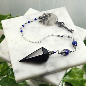 Puede incluir: Un péndulo de obsidiana negra con cadena plateada y detalles de cuentas azules. El péndulo presenta una llave decorativa plateada y un dije de laberinto plateado. El diseño está sobre una superficie de mármol blanco con follaje verde.