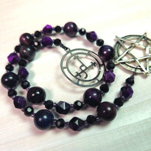 Pode incluir: Um colar de contas roxas e pretas com um amuleto de pentagrama prateado e um amuleto circular prateado com um símbolo dentro.