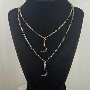 May include: Two necklaces on a black display. One necklace is gold-toned, the other is silver-toned. Each necklace features a crescent moon pendant. The chains are delicate and the pendants are small, creating an elegant look.